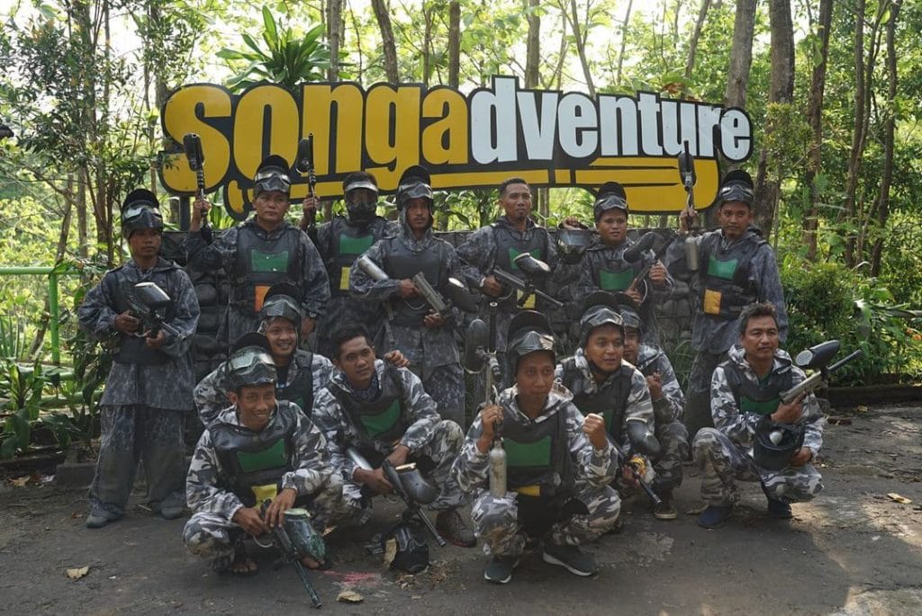 Sekelompok peserta mengenakan seragam kamuflase dan helm pelindung sedang bersiap-siap bermain paintball di area terbuka dengan rintangan buatan.
