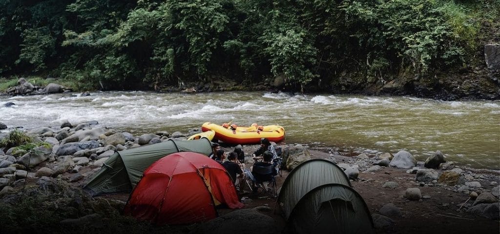 Pemandangan Alam di Songa Camp, Tenda-tenda berwarna cerah di tepi sungai dengan latar belakang hutan hijau di Songa Camp, area perkemahan yang dikelilingi alam asri.
