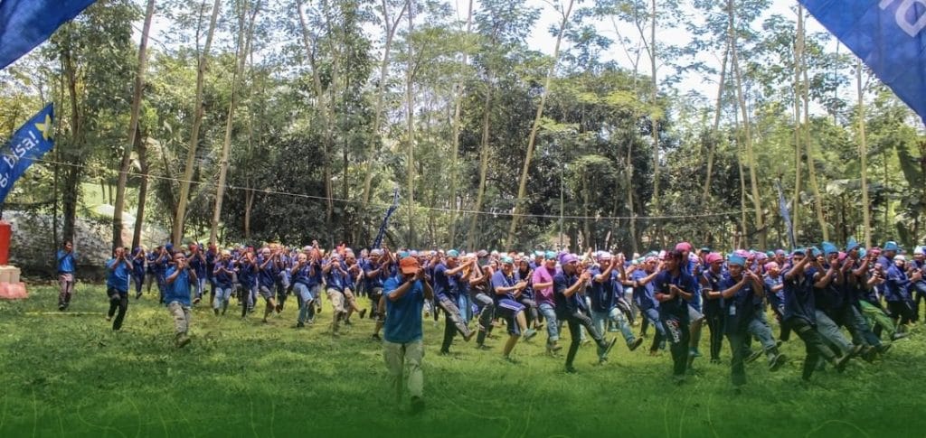 Peserta berdiri membentuk lingkaran besar di lapangan rumput, mengikuti instruksi dalam permainan ice-breaking selama sesi fun games